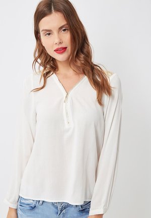 Joven con cabello largo y ondulado castaño, vistiendo una blusa blanca de mangas largas con cierre en la parte delantera y jeans de color azul claro, frente a un fondo liso.