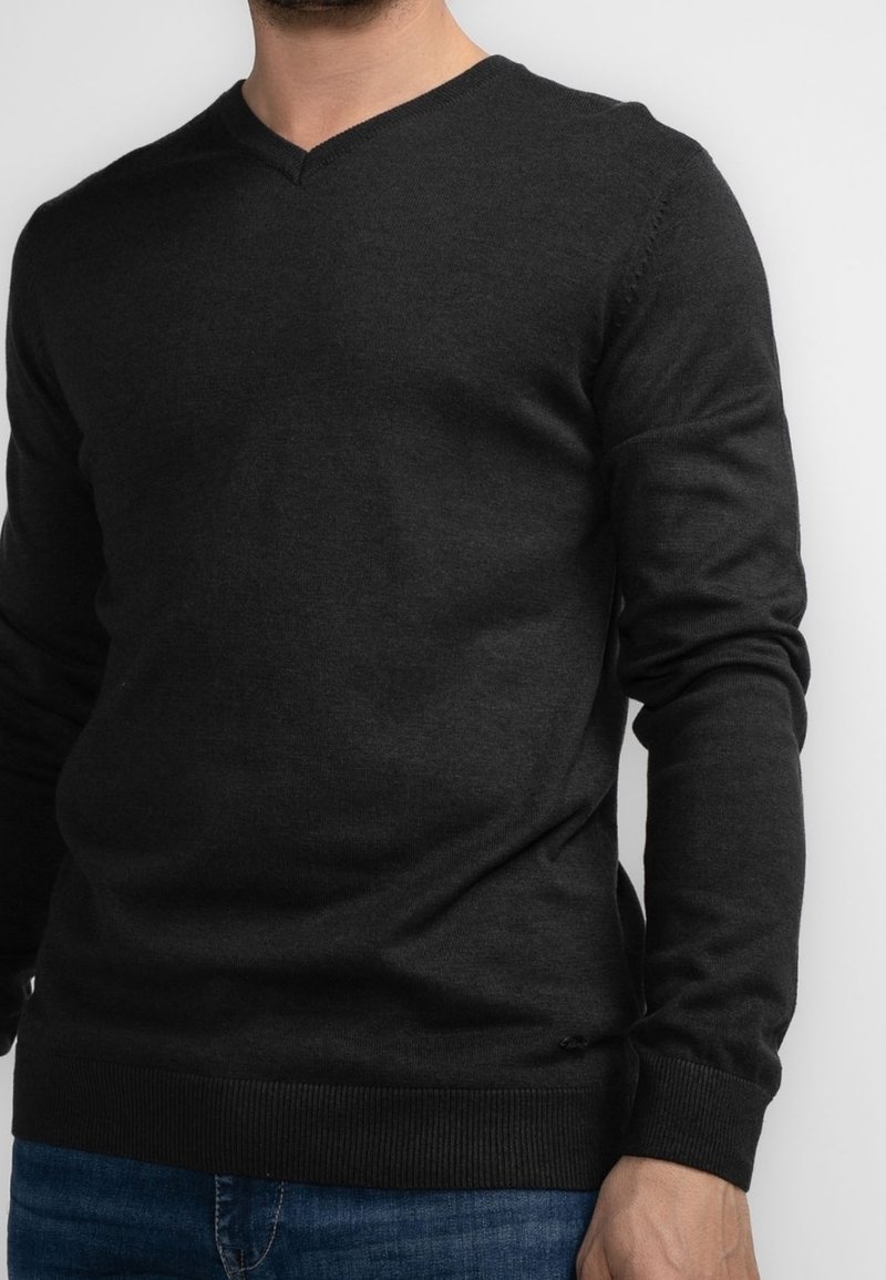 Homme portant un pull noir uni à col en V et un jean bleu, debout devant un fond blanc.