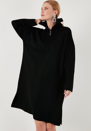 Black oversized dress with a ribbed zippered collar, featuring long sleeves and side slits for ease of movement. Soft fabric texture.
