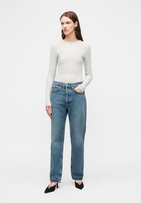 Femme debout portant un haut cintré à manches longues gris clair, un jean bleu taille haute et des escarpins noirs à bout pointu sur fond blanc.