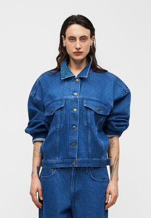 Personne aux longs cheveux noirs portant une veste en jean bleu surdimensionnée avec des boutons en métal et un pantalon en jean bleu assorti, debout devant un fond uni.