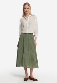 Vit långärmad skjorta med knappar ihop med en plisserad grön kjol med en subtil glans, komplementerad av ett brunt bälte och bruna skor.