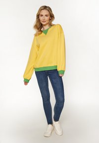 Gele oversized sweatshirt met groene accenten en geribde manchetten, gecombineerd met blauwe skinny jeans en witte sneakers. Soepele textuur.