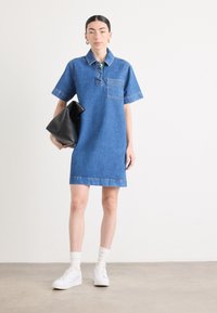 Denim skjorte dress med korte ermer, krage og frontlomme; kombinert med hvite sneakers og en svart håndveske. Flat tekstur, blå farge.