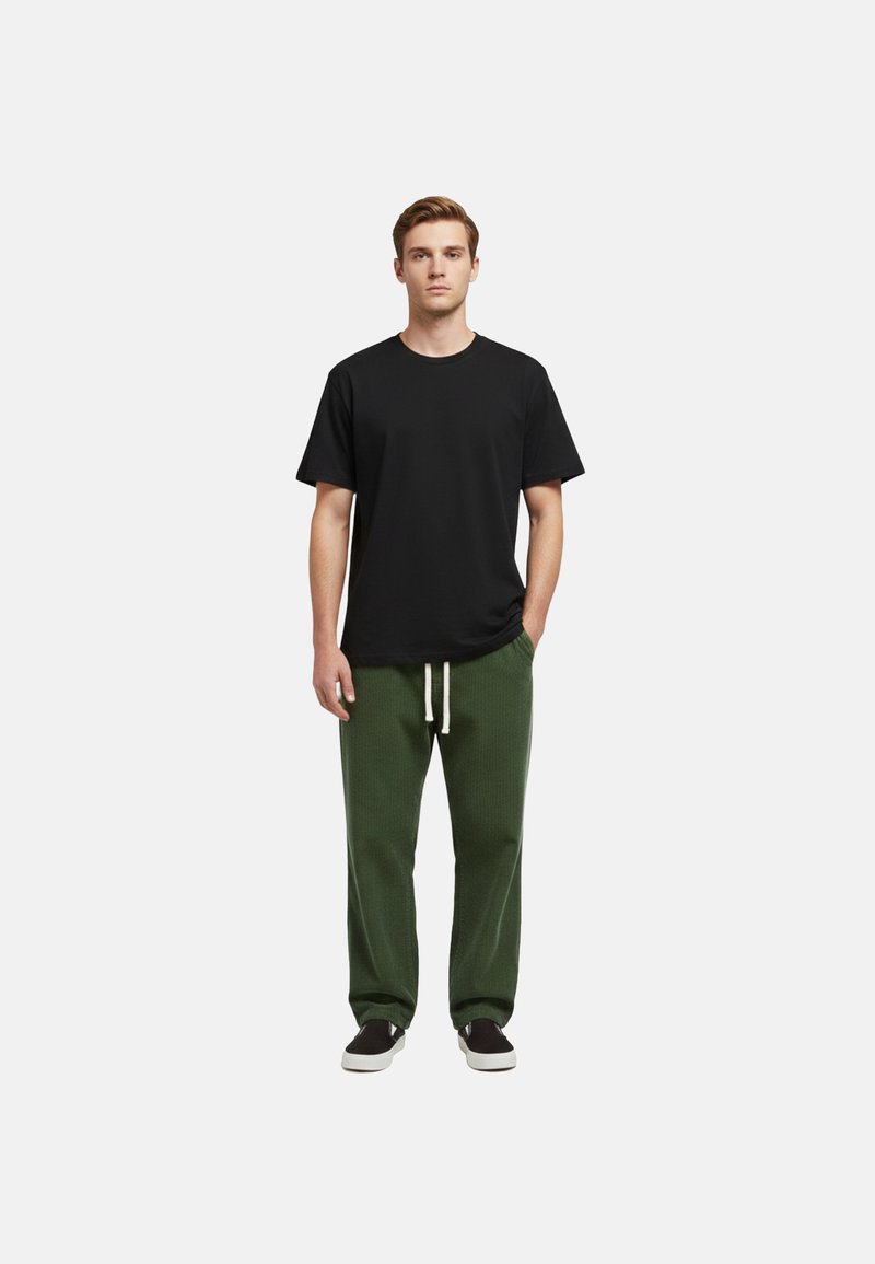 Jeune homme debout les mains dans les poches, portant un t-shirt noir, un pantalon vert avec des cordons blancs, et des chaussures noires sans lacets.