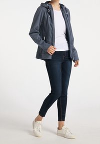 Veste à capuche gris foncé avec cordons blancs et fermetures éclair, associée à un jean foncé et des baskets blanches, modèle debout.