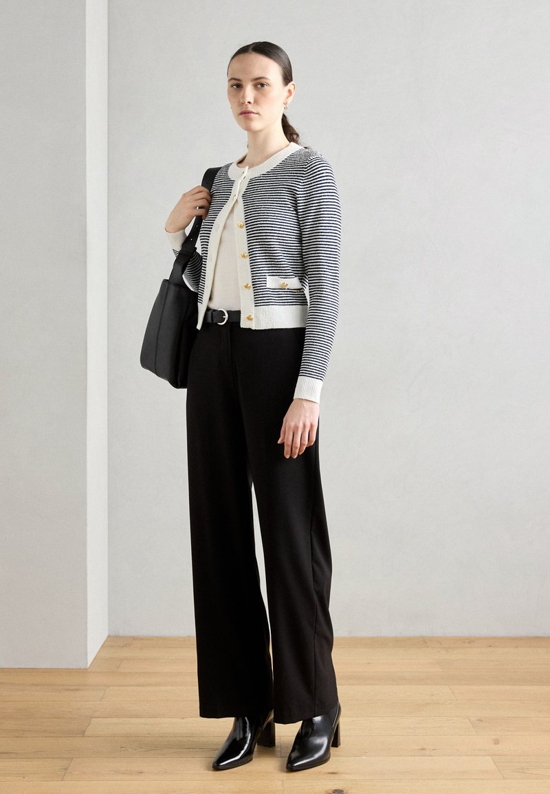 Cardigan rayé en bleu marine et blanc avec des boutons dorés, porté sur un top beige, associé à un pantalon large noir et des bottes à talons noires.
