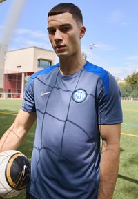 Camiseta atlética gris con acentos azules, con un escote redondo y un logotipo circular. El jugador sostiene un balón de fútbol con un patrón negro y dorado.