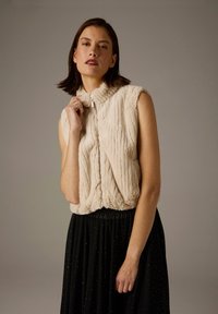 Ärmelloses, beiges Kunstpelz-Top mit Reißverschluss am Kragen, versehen mit einer vertikal gerippten Struktur. Kombiniert mit einem schwarzen, bodenlangen Rock, der mit Glitzerelementen verziert ist.