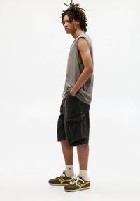 Grijze mouwloze shirt, zwarte cargoshorts met grote zakken, en groen/geel sneakers. Model staat met handen in de zakken tegen een witte achtergrond.