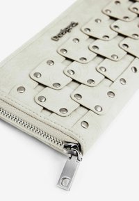 Balta piniginė, pagaminta iš tekstūruotos odos, su metaliniais kaltais ir užtrauktuku. Yra audinių dizainas su mažais stačiakampiais atvartais.