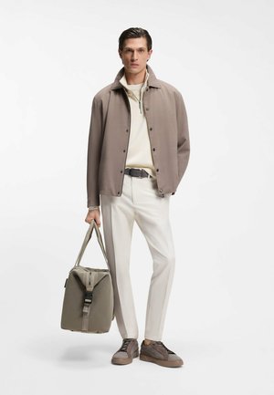 Veste extérieure marron, col roulé crème et pantalon blanc. Porte un sac vert à design structuré et des baskets marron clair avec des détails texturés.