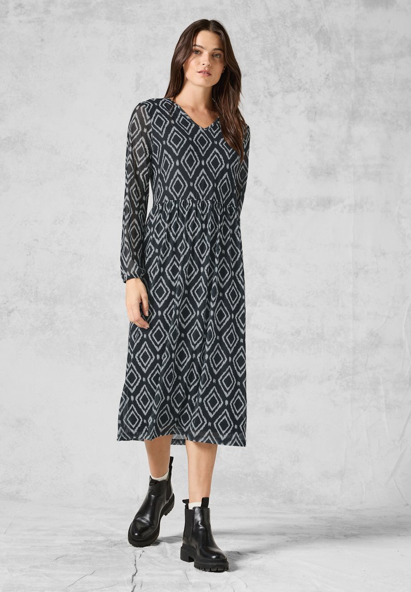Maxi-jurk met lange mouwen in zwart en grijs met diamanten patroon, V-hals, aansluitende taille en vloeiende rok; gecombineerd met zwarte enkellaarsjes.