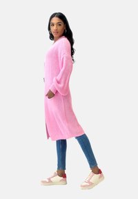 Lång, lättstickad cardigan i stark rosa, med en avslappnad passform och sidofickor, matchad med ljusblå skinny jeans och tvåfärgade sneakers.