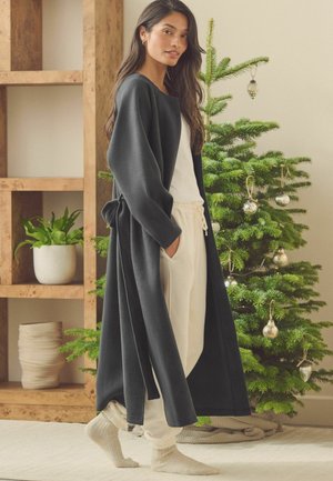 Langer dunkelgrauer Morgenmantel aus weichem Material, offene Vorderseite, mit einem Band am Bund, getragen über einem weißen Oberteil und kremfarbenen Hosen, beigen, flauschigen Socken, Weihnachtsbaum im Hintergrund.
