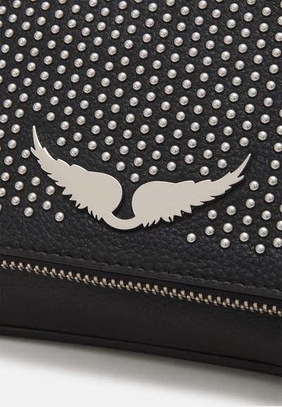 Sac en cuir noir avec des clous en argent et un emblème en forme d'aile argentée. Fermeture éclair avec détails de couture visibles.