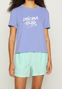 Camiseta de algodón morada con un estampado gráfico blanco, combinada con pantalones cortos verde claro. La camiseta tiene un corte relajado y mangas cortas.