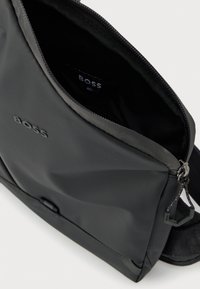 Μαύρη τσάντα ώμου με λογότυπο BOSS και μερικώς ανοιχτό φερμουάρ, που αποκαλύπτει το μαύρο εσωτερικό και μια μικρή εσωτερική ετικέτα.