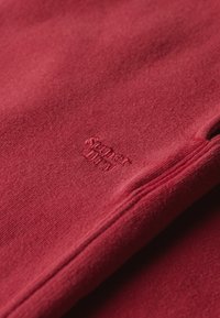 Czerwona bluza z teksturowaną tkaniną, z przeszytym logo w jaśniejszym odcieniu. Dekolt i mankiety wykończone są czystą krawędzią.