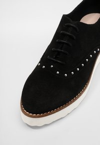 Zapato negro de ante con cordones, adornado con tachuelas plateadas, punta redonda y suela de goma blanca que presenta un detalle de costura a lo largo del borde.