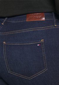 Dunkelblaue Denim-Jeans verfügen über ein Lederetikett am Bund, das mit "TOMMY HILFIGER" geprägt ist, sowie Kontrastnähte und einem Detail an der Gesäßtasche.