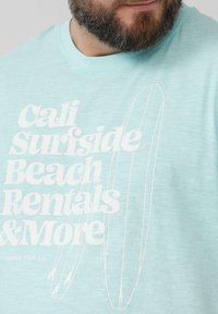 Lichtblauw T-shirt gemaakt van zachte stof met gedurfde witte tekst. Het ontwerp bevat omtrekken van surfplanken en de zin "Cali Surfside Beach Rentals & More."