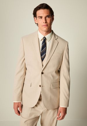 Jeune homme portant un costume beige, une chemise blanche et une cravate rayée noire et grise, debout devant un fond clair.