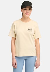 Camiseta de algodón beige con cuello redondo y mangas cortas, que presenta un pequeño logo negro de Timberland en el lado izquierdo del pecho. Combinada con leggings azul claro.