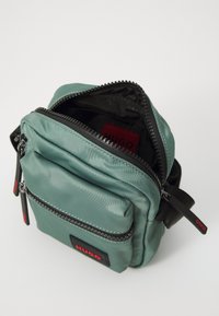 Borsa a tracolla verde in nylon con tasche con cerniera doppia, superficie testurizzata e dettaglio di branding rosso. Forma compatta e quadrata, tracolla regolabile.