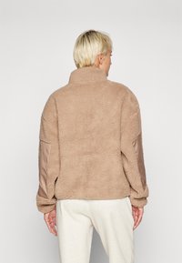 Beige fleecejakke med en blød tekstur og høj krave; har glatte satinærmer i en komplementær farve. Enkel, afslappet design.