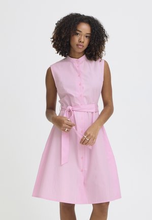 Žena s kučeravými vlasmi v rúžovom bezrukávovom šatách na gombíky viaže opasok v páse na jednoduchom pozadí.