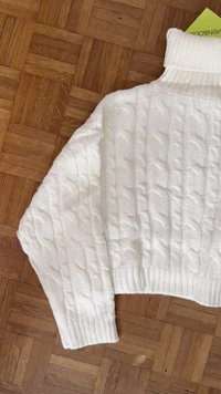 White cable-knit sweater with a high neck, cropped design, thick ribbed cuffs, and a soft texture against a wooden floor background.
