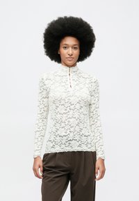 Mujer con peinado afro que lleva una blusa de encaje blanca de manga larga y pantalones sueltos de color marrón oscuro, de pie frente a un fondo claro y liso.