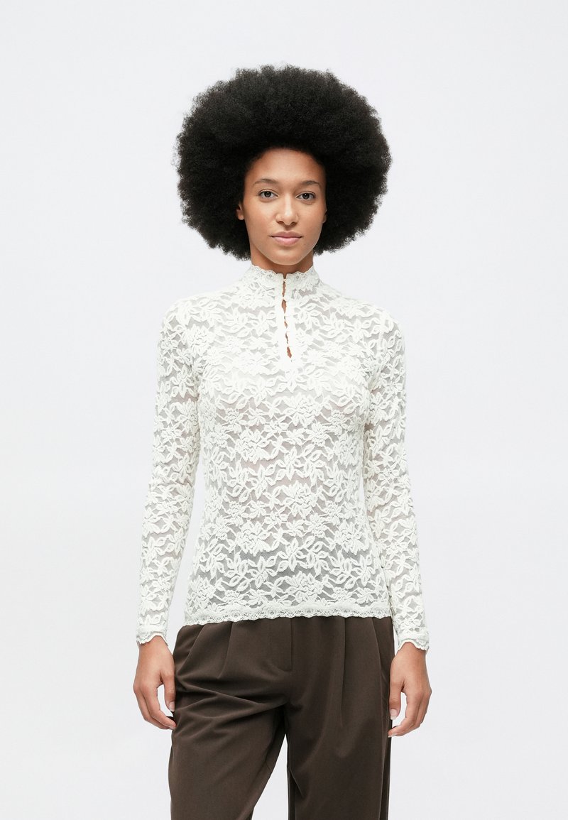 Mujer con peinado afro que lleva una blusa de encaje blanca de manga larga y pantalones sueltos de color marrón oscuro, de pie frente a un fondo claro y liso.