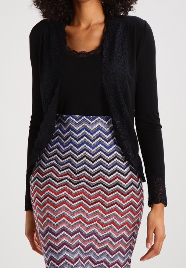 Cardigan noir avec des bordures en dentelle porté sur une jupe moulante à motifs présentant un design en zigzag multicolore dans des nuances de bleu, rouge et blanc.