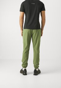 Man som bär svart kortärmad skjorta och gröna byxor med elastiska muddar, står med händerna i fickorna, vänd bort från kameran mot en vit bakgrund.