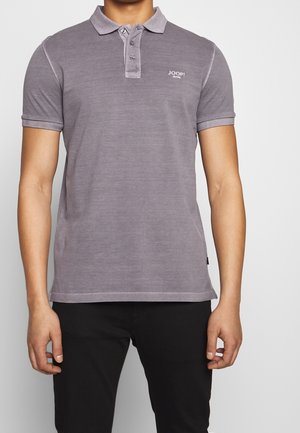 Mann trägt ein verblasstes lila kurzärmeliges Poloshirt mit dem "JOOP! JEANS"-Logo auf der Brust, kombiniert mit schwarzen Hosen, steht vor einem schlichten Hintergrund.