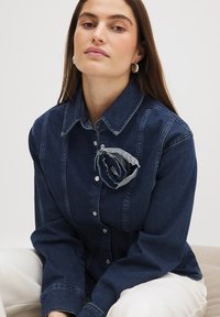 Tume sinine denim särk, millel on nööpidega kinnitamine, ja millel on rindadele kinnitatud dekoratiivne hall kangast lill. Pikad varrukad, minimalistlik disain.