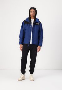 Blå vandtæt jakke med sorte detaljer, trykknapper foran, justerbar hætte, parret med sorte bukser og crèmefarvede sneakers.