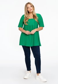 Groene tuniektop met een vierkante halslijn, korte mouwen en een smockbuste, gecombineerd met donkere jeans en witte sneakers. Glad stofoppervlak.