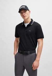 Sort polo-shirt med hvide striber på kraven, med et logo på brystet. Grå bukser og et sort bælte fuldender outfittet.