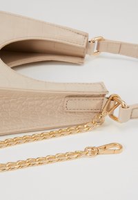 Beżowa torebka o wzorze krokodyla z złotym łańcuszkiem, charakteryzująca się gładką teksturą i zaokrąglonym kształtem. Posiada klips do przypięcia paska.