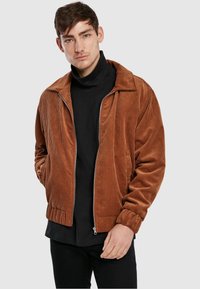 Brown corduroy zip-up jacket with a pointed collar, ribbed cuffs, and waistband, layered over a black turtleneck. Smooth texture.