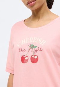 Ροζ βαμβακερό μπλουζάκι με φαρδύ λαιμόκοψη, διακοσμημένο με γραφικό εκτύπωμα κόκκινων κερασιών και κείμενο «CHERRISH the Night» σε αντίθετα χρώματα.