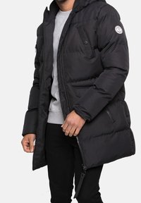 Veste matelassée noire avec capuche, fermeture éclair sur le devant, poche zippée sur le côté, et écusson rond avec logo sur la manche gauche. Associée à une chemise grise.