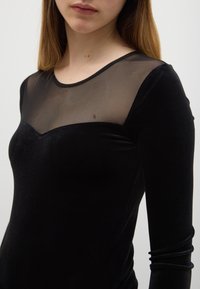 Zwarte top met lange mouwen en een aansluitend model. Voorzien van een transparant mesh inzetstuk bij de halslijn en een zachte, gladde fluwelen textuur.