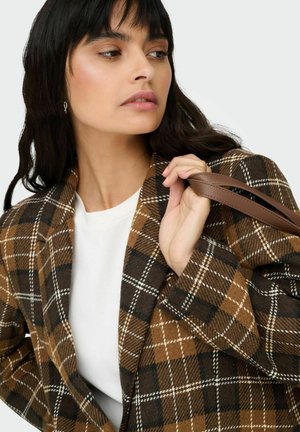 Rudas languotas švarkas iš vilnos, su baltomis ir juodomis linijomis, dėvimas ant balto viršaus, rankos laiko rudos odos dirželį.