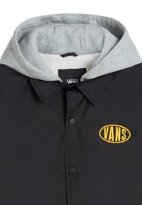 Giacca nera con cappuccio foderato in pile grigio. Presenta bottoni a pressione e un logo circolare giallo Vans sulla parte sinistra del petto.