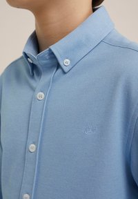 Polo de culoare albastru deschis, realizat dintr-un material texturat, cu guler, nasturi albi și un mic logo brodat pe partea stângă.