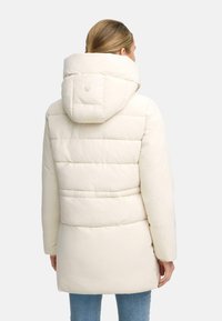 Veste matelassée de couleur crème avec capuche, présentant une texture matelassée, des coutures horizontales et un détail de logo au dos.